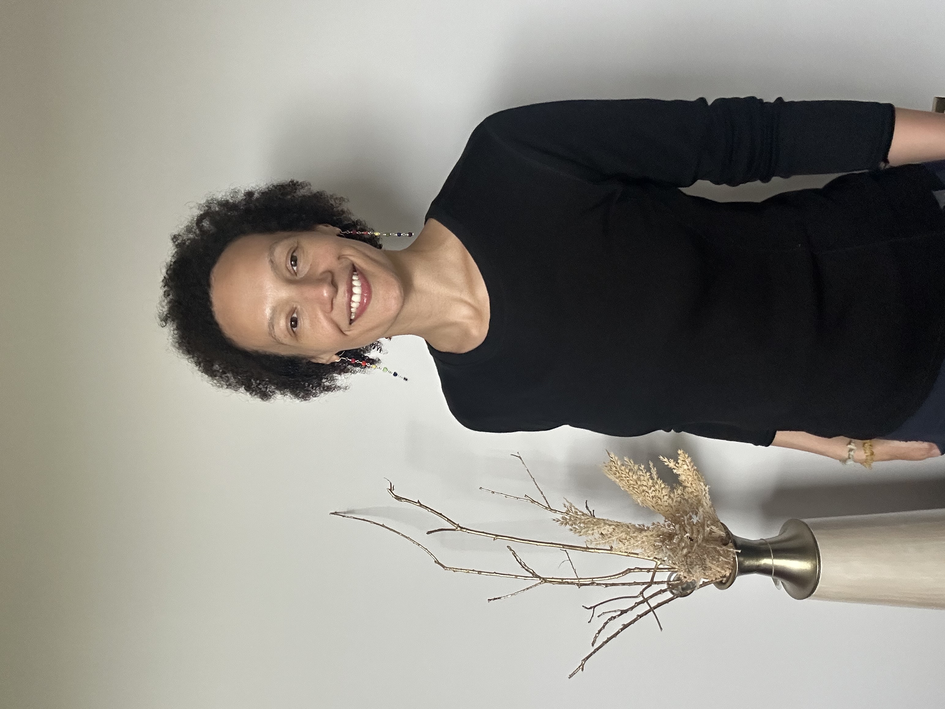 Portrait of Living Well Studios Practioner Bree, LMT stands in front of a neutral wall with her natural afro and smiling at the camera 