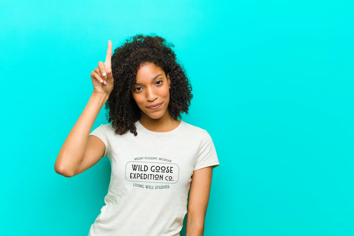 A smiling Black woman with natural hair pointing upward while wearing a Living Well Studios original logo T-shirt against a bright teal background